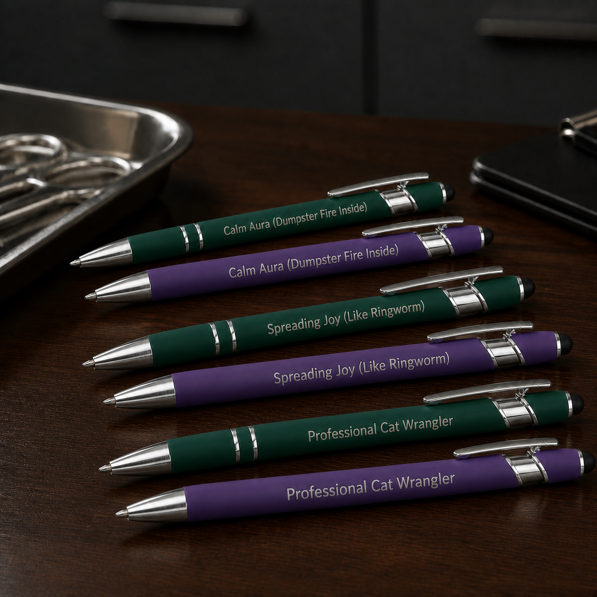 Six laser-engraved veterinary pens in green and purple with stainless steel hardware on a dark clinic desk